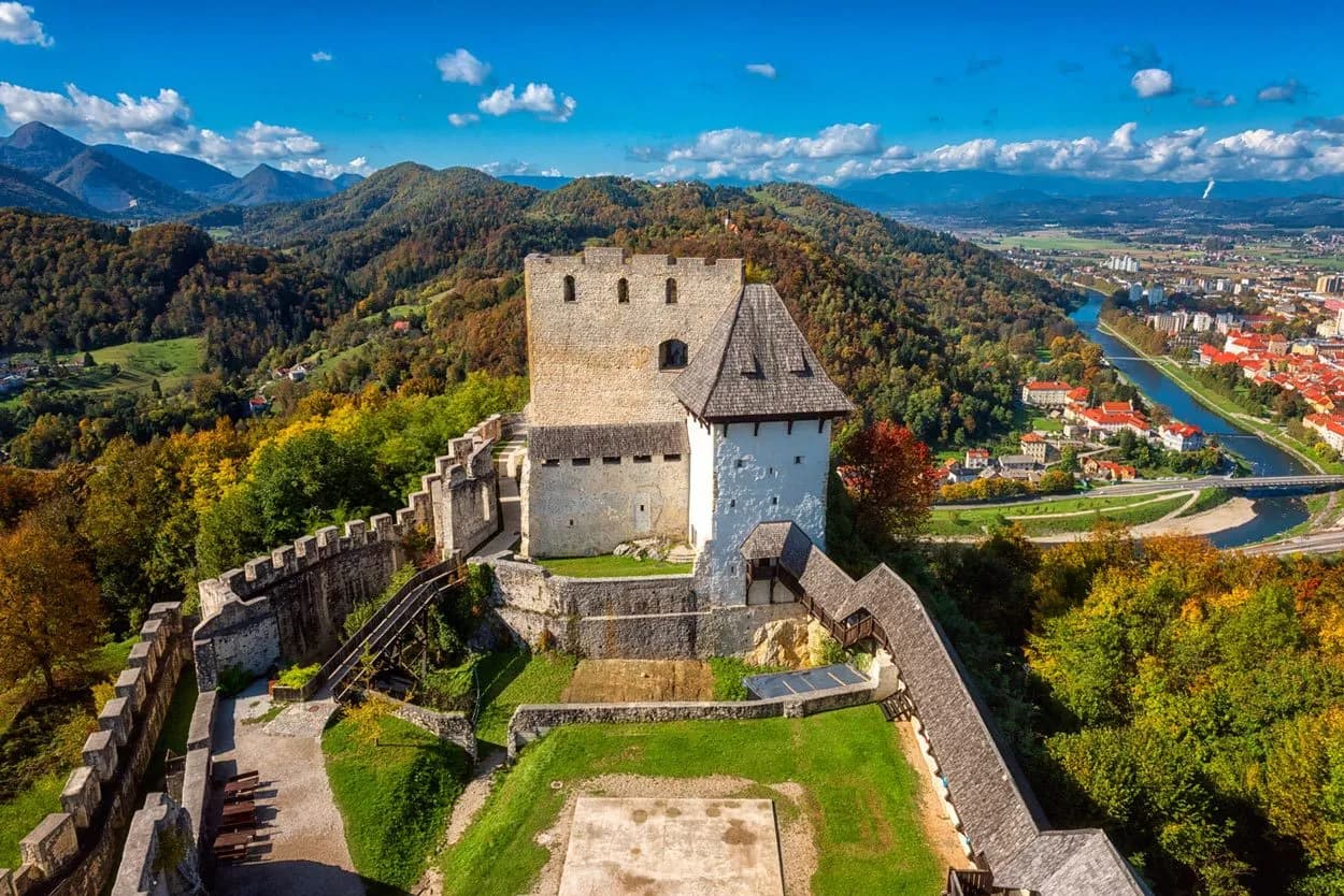 Historic castle ruins overlooking a city and river valley with autumn foliage under blue sky.